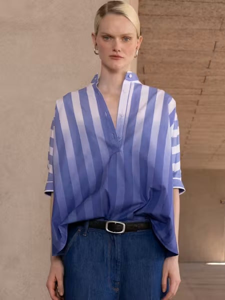 Someone stands indoors in the NUDE - OMBRE STRIPE OVERSIZED SHIRT by NUDE, featuring blue-and-white vertical stripes, a V-neck, and short sleeves. They pair it with blue jeans and a black belt against neutral-colored walls and ceiling.