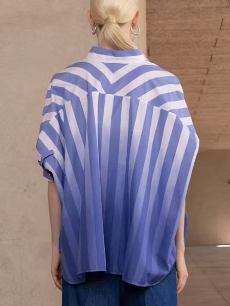 Someone stands indoors in the NUDE - OMBRE STRIPE OVERSIZED SHIRT by NUDE, featuring blue-and-white vertical stripes, a V-neck, and short sleeves. They pair it with blue jeans and a black belt against neutral-colored walls and ceiling.