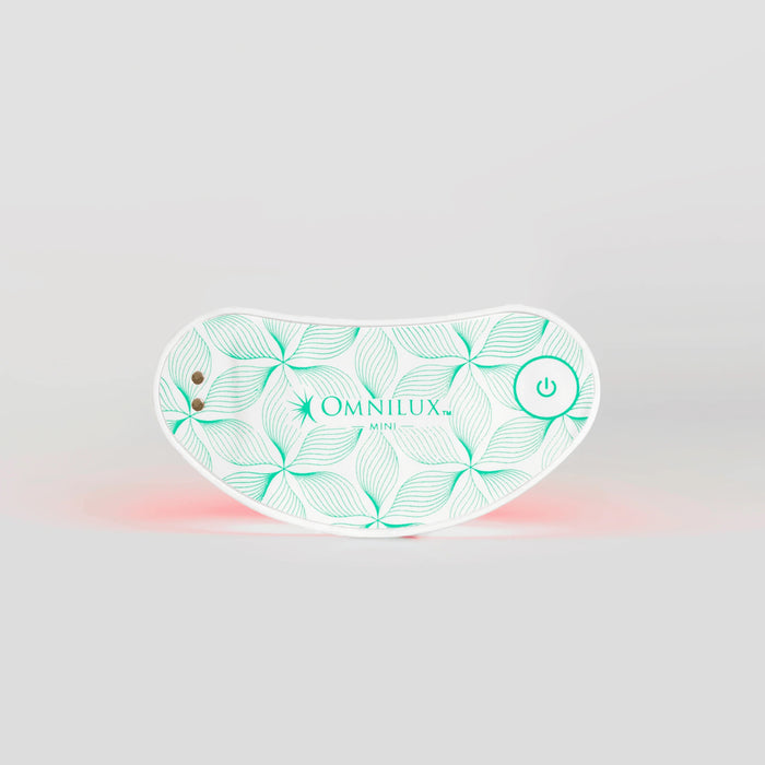 White wearable eye mask with a green leaf pattern, labeled “OMNILUX – EYE BRIGHTENER KIT,” featuring a power button and two small metal contacts, softly glowing with a red light underneath to deliver targeted red light therapy for brightening the eye area against a plain gray background.