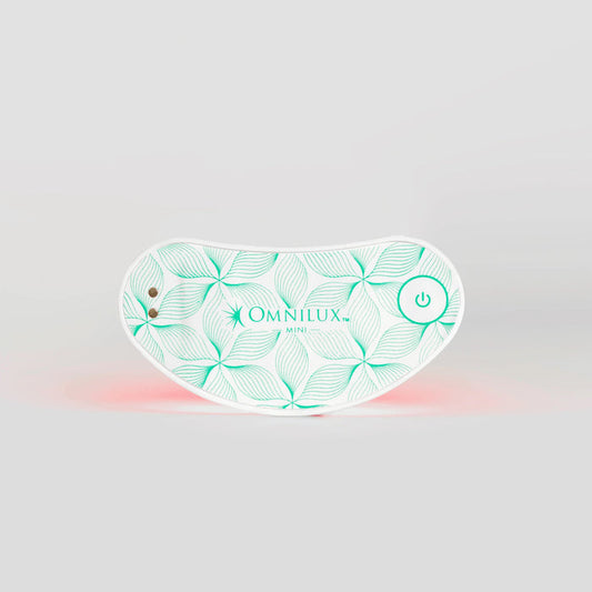 White wearable eye mask with a green leaf pattern, labeled “OMNILUX – EYE BRIGHTENER KIT,” featuring a power button and two small metal contacts, softly glowing with a red light underneath to deliver targeted red light therapy for brightening the eye area against a plain gray background.