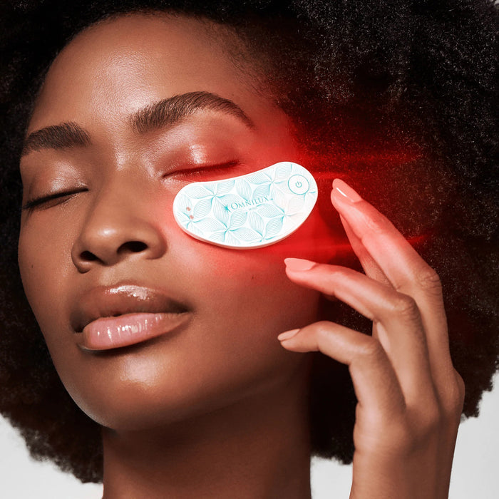 White wearable eye mask with a green leaf pattern, labeled “OMNILUX – EYE BRIGHTENER KIT,” featuring a power button and two small metal contacts, softly glowing with a red light underneath to deliver targeted red light therapy for brightening the eye area against a plain gray background.