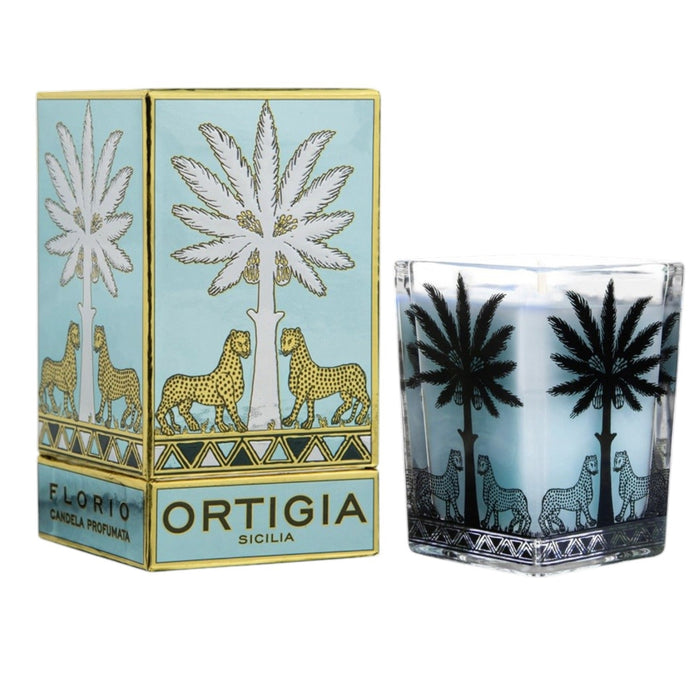 Four ORTIGIA SICILIA - FLORIO LARGE SQUARE CANDLES by ORTIGIA SICILIA, each in a decorative patterned glass holder featuring palm tree and animal designs, are displayed alongside their matching boxes on a marble surface. The pale blue wax of the candles complements the vibrant arrangement of colorful tulips, Sicilian spring flowers, and other blooms surrounding them.