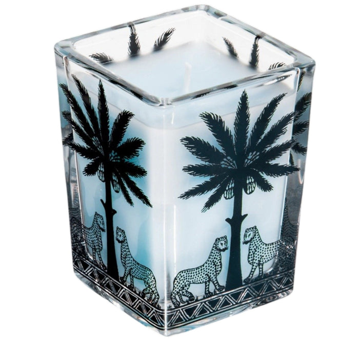 Four ORTIGIA SICILIA - FLORIO LARGE SQUARE CANDLES by ORTIGIA SICILIA, each in a decorative patterned glass holder featuring palm tree and animal designs, are displayed alongside their matching boxes on a marble surface. The pale blue wax of the candles complements the vibrant arrangement of colorful tulips, Sicilian spring flowers, and other blooms surrounding them.