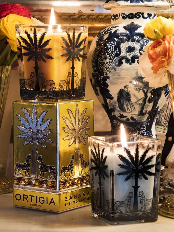 Four ORTIGIA SICILIA - FLORIO LARGE SQUARE CANDLES by ORTIGIA SICILIA, each in a decorative patterned glass holder featuring palm tree and animal designs, are displayed alongside their matching boxes on a marble surface. The pale blue wax of the candles complements the vibrant arrangement of colorful tulips, Sicilian spring flowers, and other blooms surrounding them.