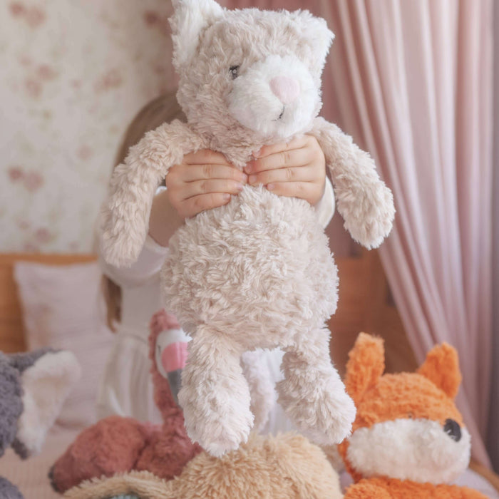 Stack of plush toys including a cat, bear, and fox on a bed with a soft pink curtain in the background.