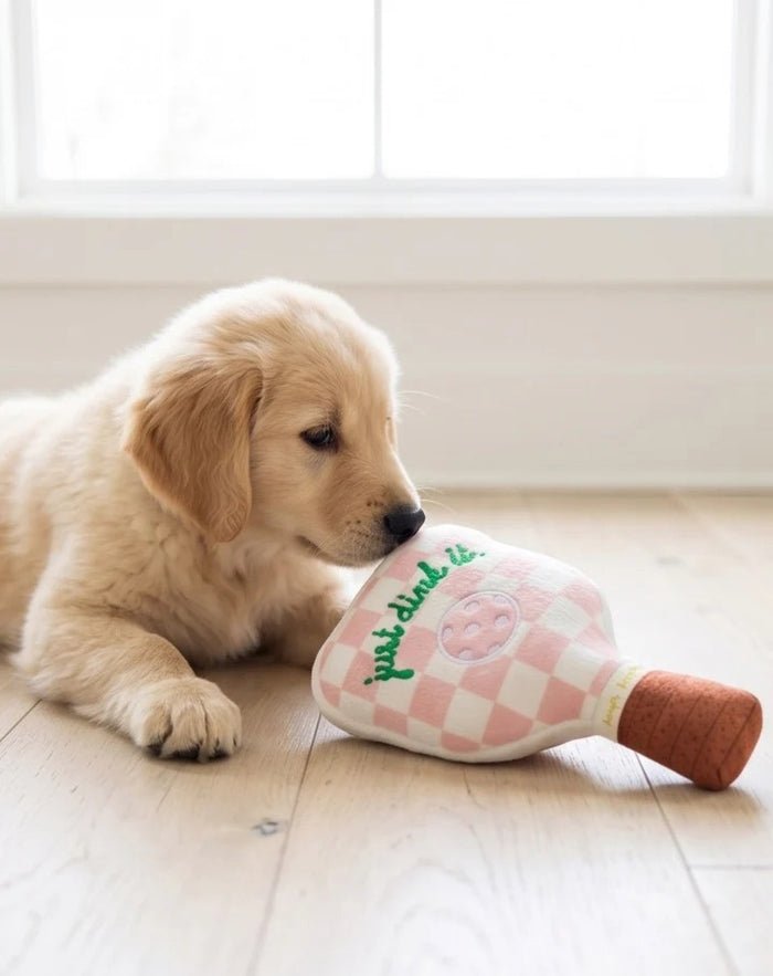 The HAUTE DIGGITY DOG PINK CHECKER PICKLEBALL PADDLE DOG TOY is a plush, paddle-shaped squeaky dog toy featuring a fun pink checkered design, an embroidered pickleball, the phrase "just dink it." in green, and a brown handle—perfect for pups who want to join in on your pickleball passion.