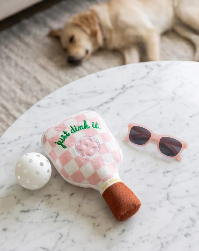 The HAUTE DIGGITY DOG PINK CHECKER PICKLEBALL PADDLE DOG TOY is a plush, paddle-shaped squeaky dog toy featuring a fun pink checkered design, an embroidered pickleball, the phrase "just dink it." in green, and a brown handle—perfect for pups who want to join in on your pickleball passion.