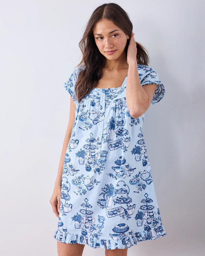 A woman stands barefoot on a wood floor, wearing the PRINTFRESH High Tea Toile Pintuck Nightgown in Notting Hill Blue—a vintage-inspired piece with short sleeves, a ruffled hem, and a charming blue illustrated print. She poses against a white wall with a relaxed smile.