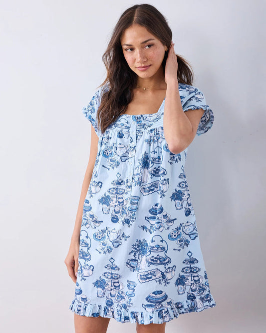 A woman stands barefoot on a wood floor, wearing the PRINTFRESH High Tea Toile Pintuck Nightgown in Notting Hill Blue—a vintage-inspired piece with short sleeves, a ruffled hem, and a charming blue illustrated print. She poses against a white wall with a relaxed smile.