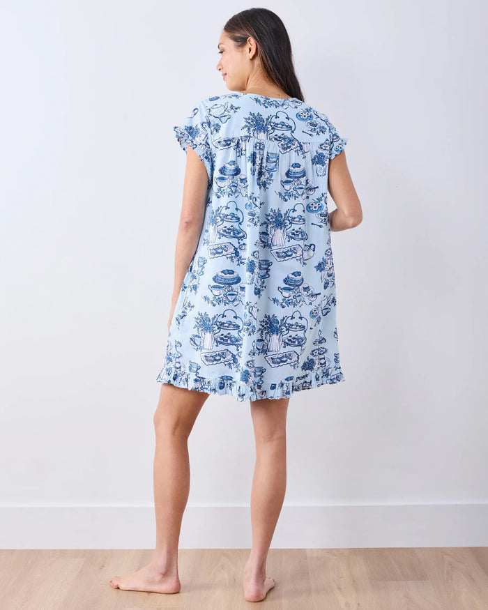 A woman wearing the PRINTFRESH - High Tea Toile Pintuck Nightgown in Notting Hill Blue, featuring blue dessert and tea set illustrations, stands against a plain light-colored wall, touching her hair and gazing slightly to the side.