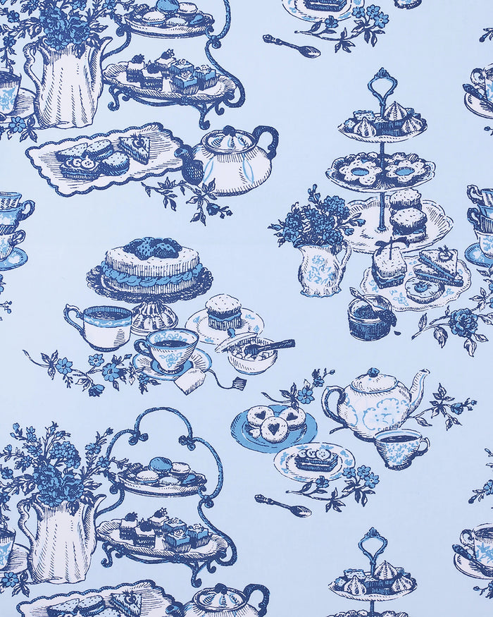 A woman stands barefoot on a wood floor, wearing the PRINTFRESH High Tea Toile Pintuck Nightgown in Notting Hill Blue—a vintage-inspired piece with short sleeves, a ruffled hem, and a charming blue illustrated print. She poses against a white wall with a relaxed smile.