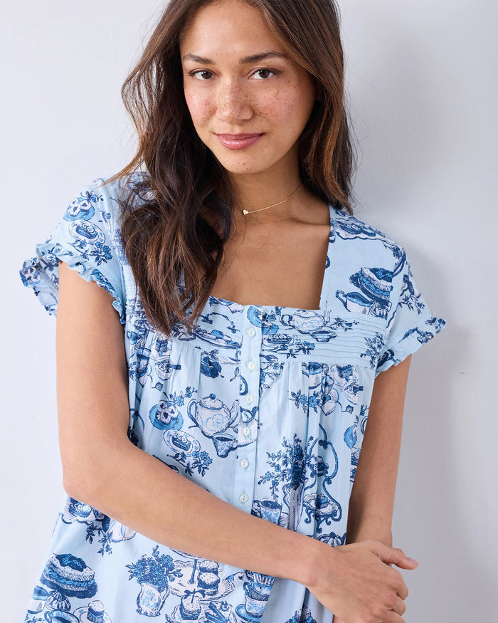 A woman wearing the PRINTFRESH - High Tea Toile Pintuck Nightgown in Notting Hill Blue, featuring blue dessert and tea set illustrations, stands against a plain light-colored wall, touching her hair and gazing slightly to the side.