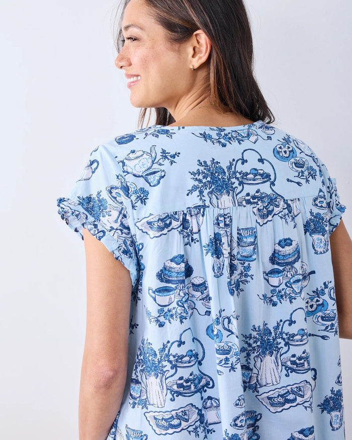 A woman stands barefoot on a wood floor, wearing the PRINTFRESH High Tea Toile Pintuck Nightgown in Notting Hill Blue—a vintage-inspired piece with short sleeves, a ruffled hem, and a charming blue illustrated print. She poses against a white wall with a relaxed smile.