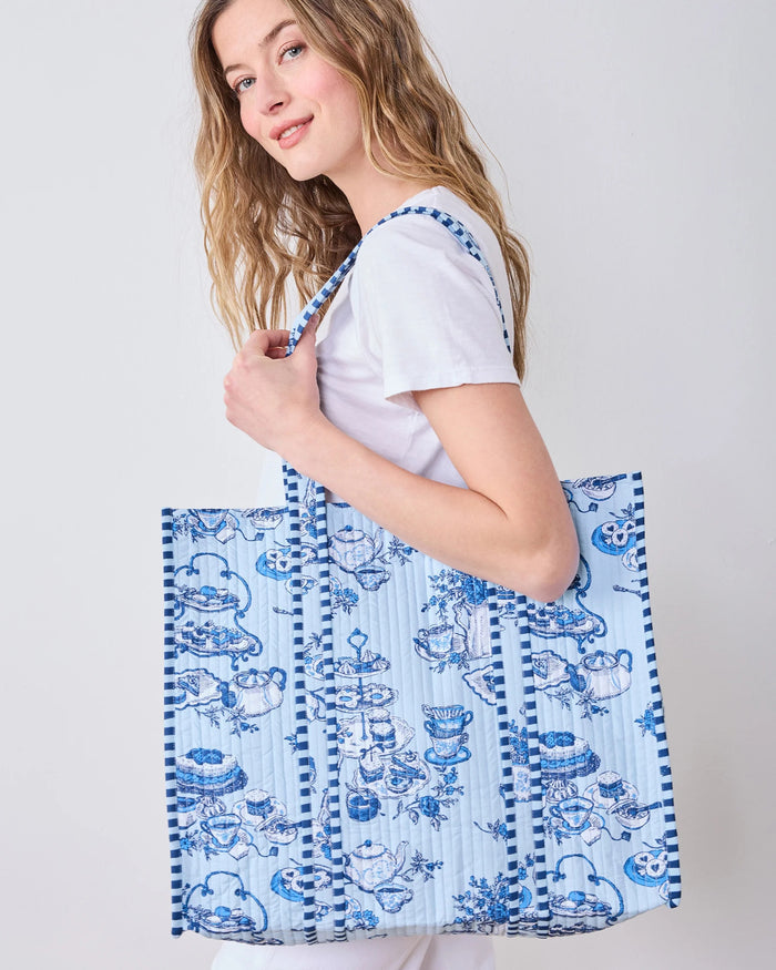 Smiling woman in a white shirt carries the spacious PRINTFRESH - HIGH TEA TOILE TRAVELER TOTE, featuring a bold blue-and-white pattern and draped over her shoulder as she stands against a plain light background—the perfect organic cotton tote bag from PRINTFRESH for the tea lover gift in your life.