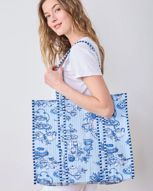 Smiling woman in a white shirt carries the spacious PRINTFRESH - HIGH TEA TOILE TRAVELER TOTE, featuring a bold blue-and-white pattern and draped over her shoulder as she stands against a plain light background—the perfect organic cotton tote bag from PRINTFRESH for the tea lover gift in your life.