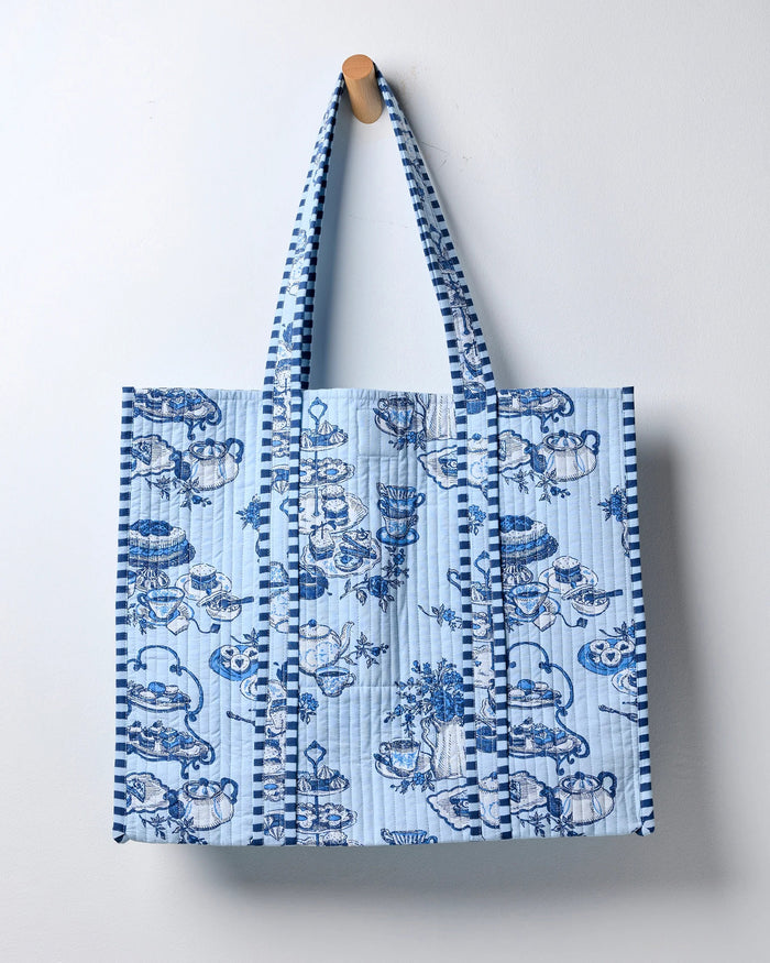 Smiling woman in a white shirt carries the spacious PRINTFRESH - HIGH TEA TOILE TRAVELER TOTE, featuring a bold blue-and-white pattern and draped over her shoulder as she stands against a plain light background—the perfect organic cotton tote bag from PRINTFRESH for the tea lover gift in your life.
