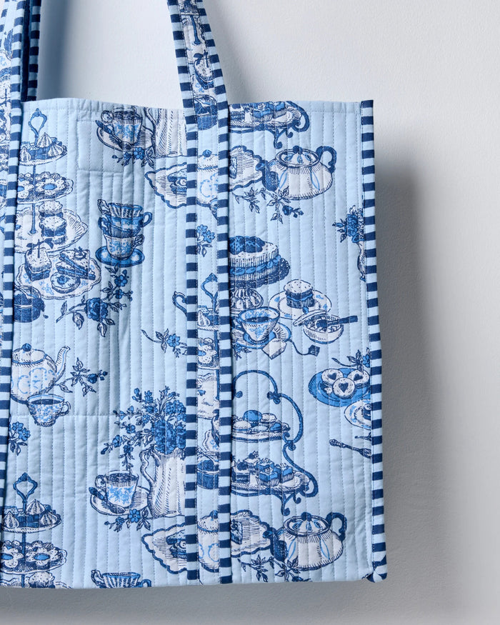 Smiling woman in a white shirt carries the spacious PRINTFRESH - HIGH TEA TOILE TRAVELER TOTE, featuring a bold blue-and-white pattern and draped over her shoulder as she stands against a plain light background—the perfect organic cotton tote bag from PRINTFRESH for the tea lover gift in your life.