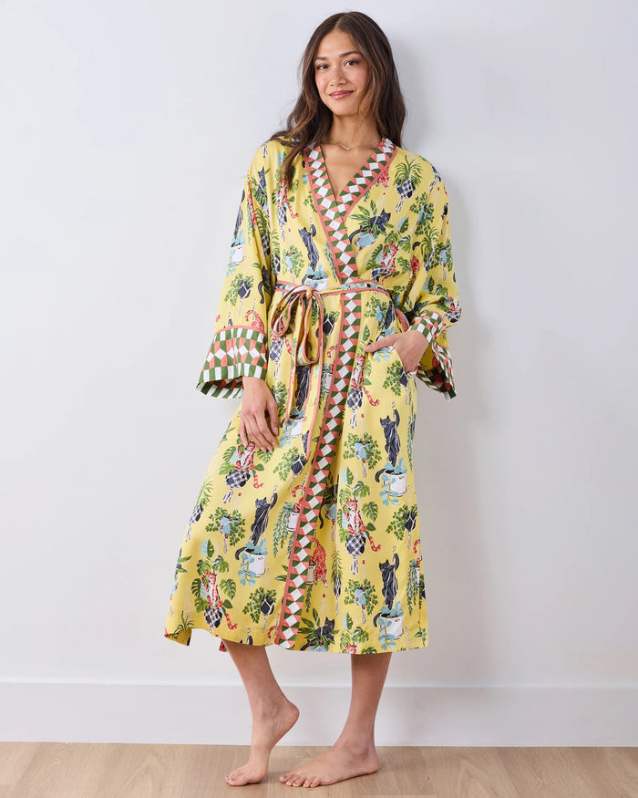 A woman stands barefoot against a white wall, wearing the PRINTFRESH - Macrame Mischief Eco Satin Robe in Sunshine—a long yellow robe featuring colorful animal and botanical designs, geometric trim, and a matching belt—perfect for any cat lover.