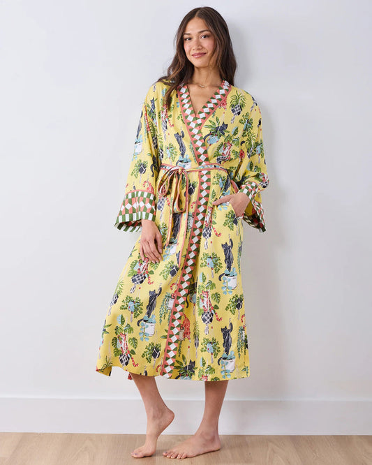 A woman stands barefoot against a white wall, wearing the PRINTFRESH - Macrame Mischief Eco Satin Robe in Sunshine—a long yellow robe featuring colorful animal and botanical designs, geometric trim, and a matching belt—perfect for any cat lover.