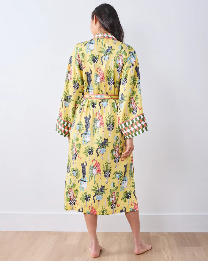 A woman stands barefoot against a white wall, wearing the PRINTFRESH - Macrame Mischief Eco Satin Robe in Sunshine—a long yellow robe featuring colorful animal and botanical designs, geometric trim, and a matching belt—perfect for any cat lover.