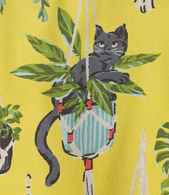 A woman stands barefoot against a white wall, wearing the PRINTFRESH - Macrame Mischief Eco Satin Robe in Sunshine—a long yellow robe featuring colorful animal and botanical designs, geometric trim, and a matching belt—perfect for any cat lover.