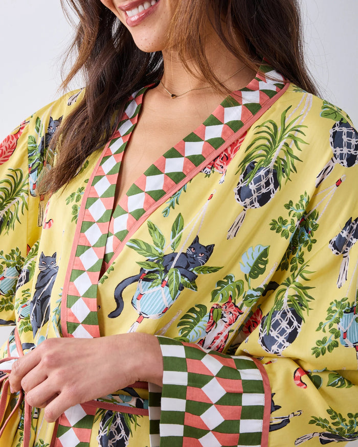 A woman stands barefoot against a white wall, wearing the PRINTFRESH - Macrame Mischief Eco Satin Robe in Sunshine—a long yellow robe featuring colorful animal and botanical designs, geometric trim, and a matching belt—perfect for any cat lover.