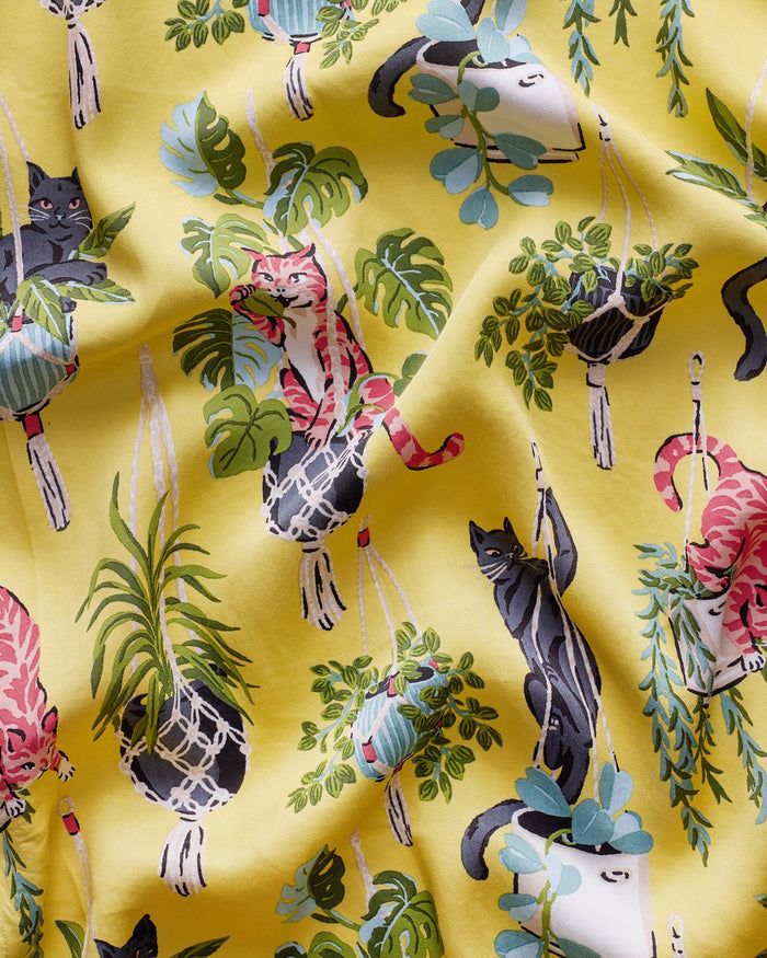 A woman stands barefoot against a white wall, wearing the PRINTFRESH - Macrame Mischief Eco Satin Robe in Sunshine—a long yellow robe featuring colorful animal and botanical designs, geometric trim, and a matching belt—perfect for any cat lover.