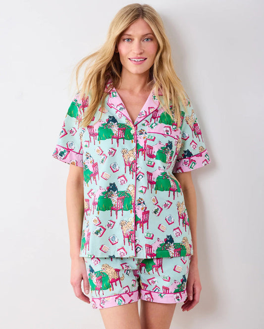Dressed in PRINTFRESH’s Mahjong Menagerie Pajama Short Set, this person sports matching pastel organic cotton sleepwear featuring a playful cat print with tables, books, and chairs. The set includes a short-sleeve button-up shirt and shorts, both finished with light pink trim.
