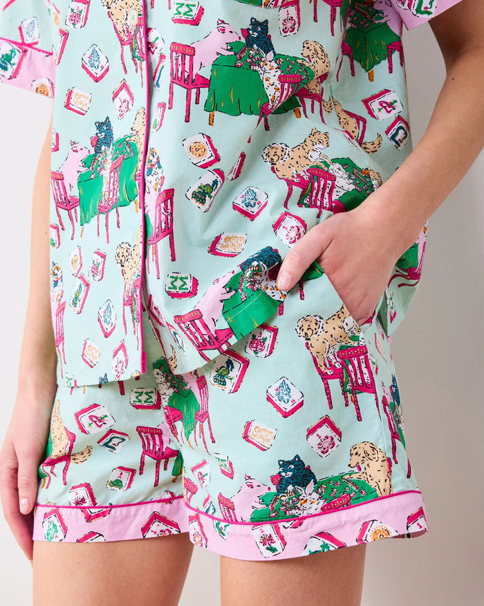 A woman with long blonde hair smiles as she stands against a plain white background, wearing the PRINTFRESH - MAHJONG MENAGERIE PAJAMA SHORT SET by PRINTFRESH, a matching short-sleeve button-up pajama top and shorts hand screen-printed with colorful cat and chair patterns.
