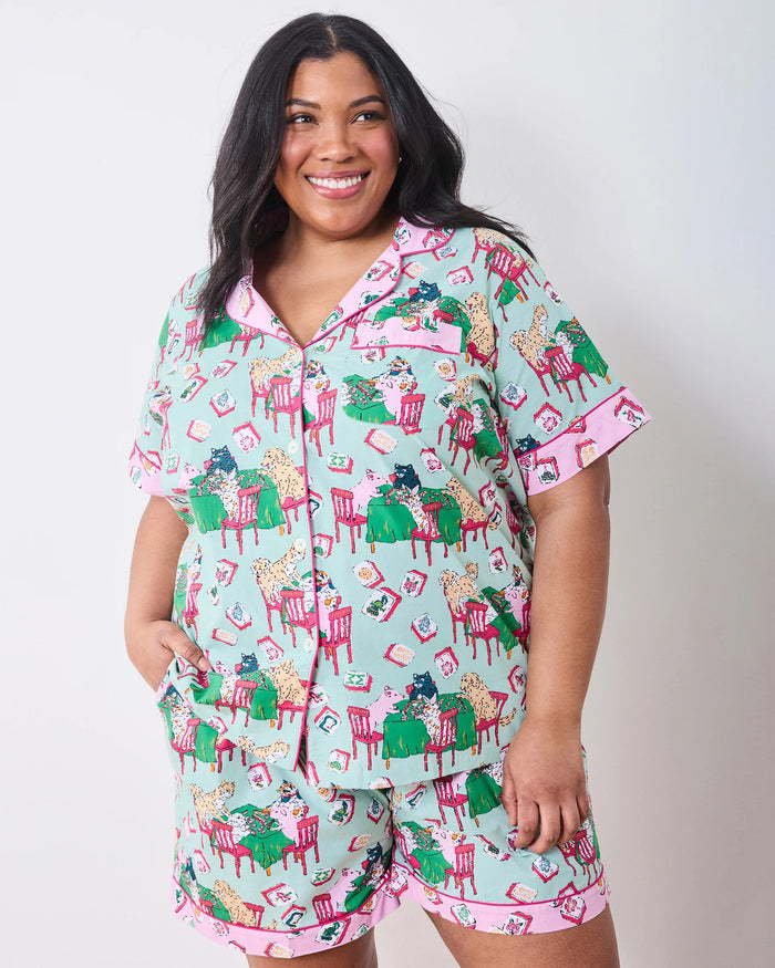 Dressed in PRINTFRESH’s Mahjong Menagerie Pajama Short Set, this person sports matching pastel organic cotton sleepwear featuring a playful cat print with tables, books, and chairs. The set includes a short-sleeve button-up shirt and shorts, both finished with light pink trim.