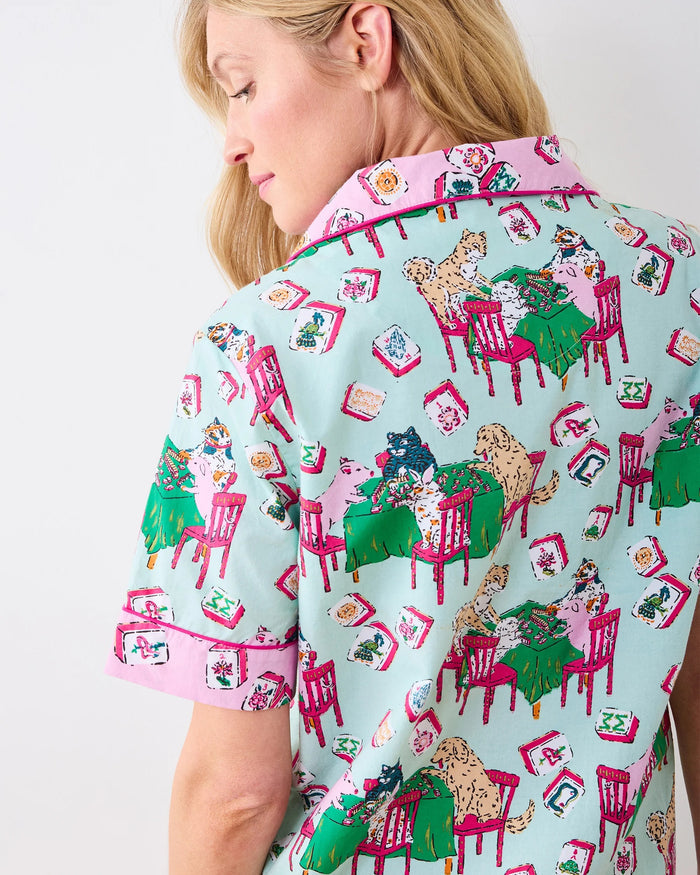 A woman with long blonde hair smiles as she stands against a plain white background, wearing the PRINTFRESH - MAHJONG MENAGERIE PAJAMA SHORT SET by PRINTFRESH, a matching short-sleeve button-up pajama top and shorts hand screen-printed with colorful cat and chair patterns.