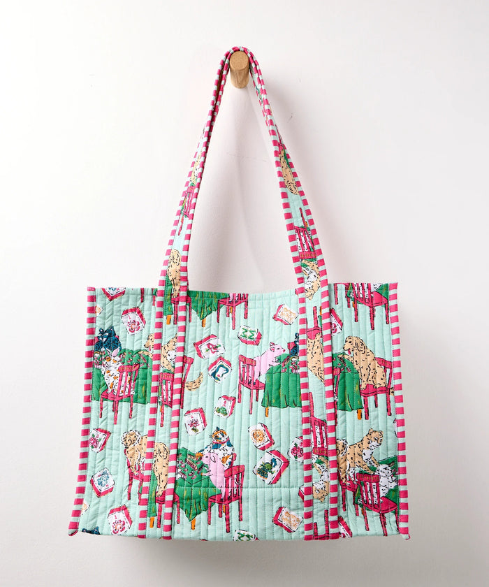 A woman in a white shirt and white pants smiles while holding the PRINTFRESH Mahjong Menagerie Traveler Tote—an eye-catching, quilted bag with colorful red, green, and blue designs—over her shoulder, standing against a plain light-colored background.
