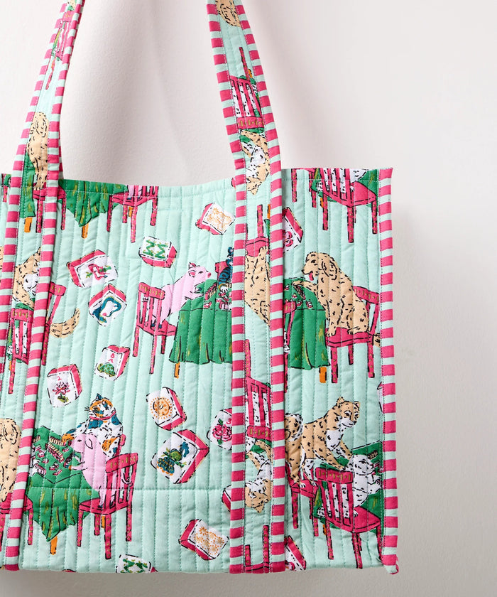 A woman in a white shirt and white pants smiles while holding the PRINTFRESH Mahjong Menagerie Traveler Tote—an eye-catching, quilted bag with colorful red, green, and blue designs—over her shoulder, standing against a plain light-colored background.