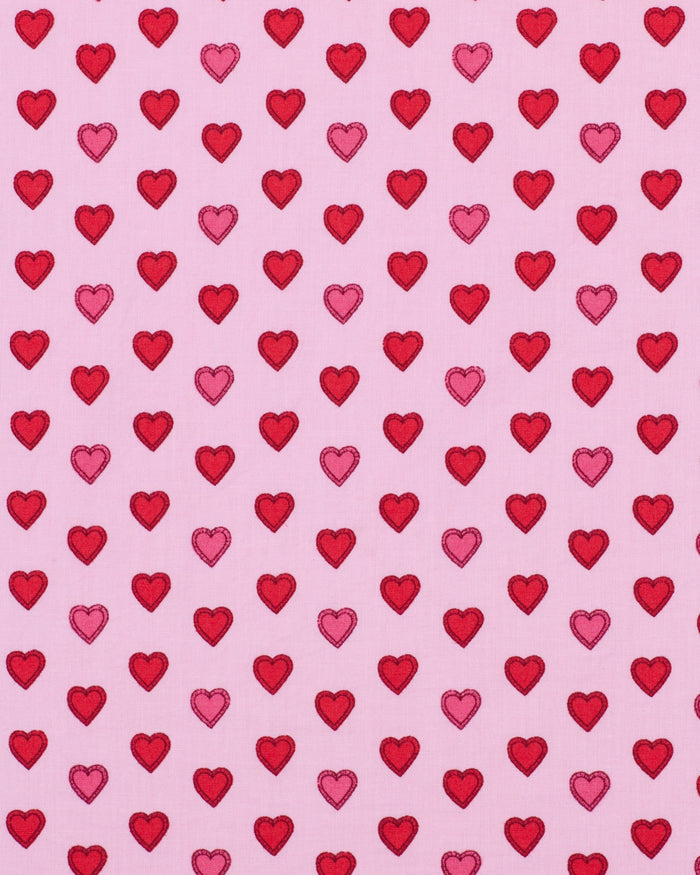 A woman stands barefoot on a wooden floor, wearing the PRINTFRESH - MINI CANDY HEARTS LONG PAJAMA SET by PRINTFRESH. The pajamas feature pink fabric decorated with a red heart pattern. She has long blonde hair and a relaxed expression, posing against a plain white wall.