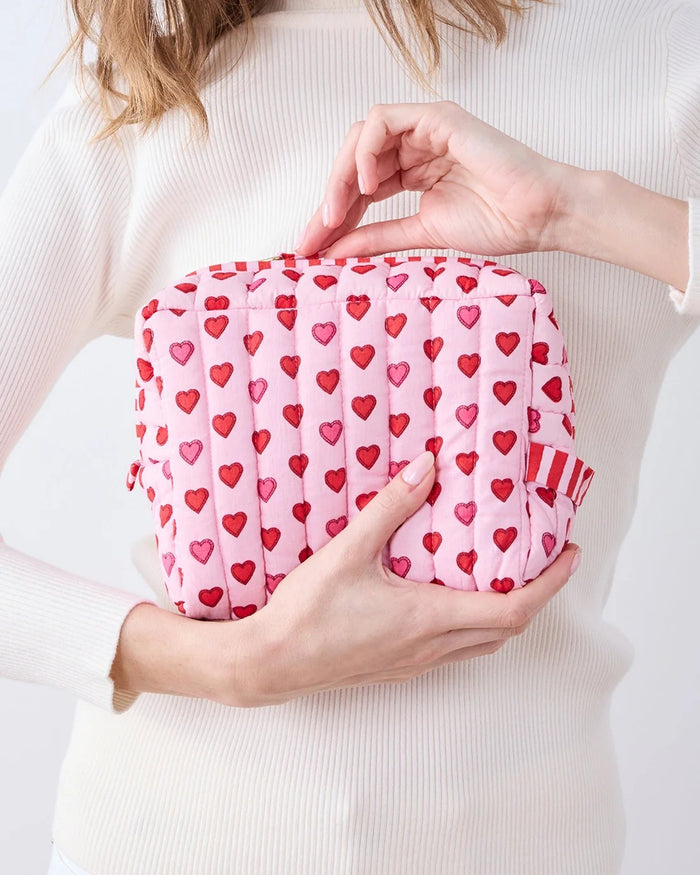 A person in a white ribbed sweater holds one of the charming heart pouches from the PRINTFRESH Mini Candy Hearts Three Pouch Set—a quilted cotton pouch from PRINTFRESH, finished in pink and adorned with red and pink hearts, plus a red-and-white striped tab.