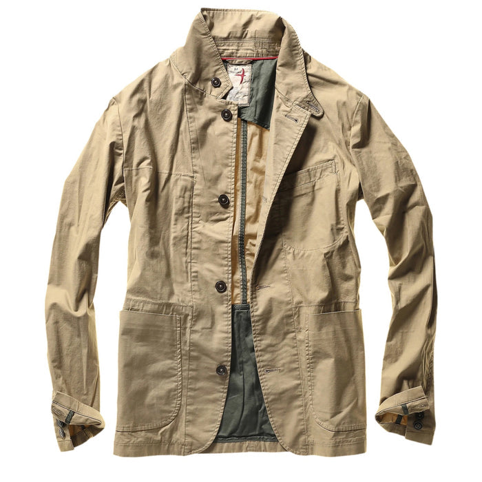 A person wears the RELWEN Flyweight Flex Blazer in British Khaki over a blue and white checkered shirt with green pants, standing hands-in-pockets against a white background. Face not visible.