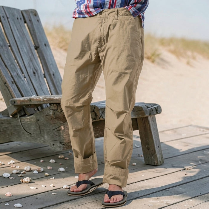 Wearing RELWEN's Flyweight Flex Chino in British Khaki, a person stands on a beachside boardwalk near a weathered bench, with flip-flops and a plaid shirt, seashells and sand in the background.