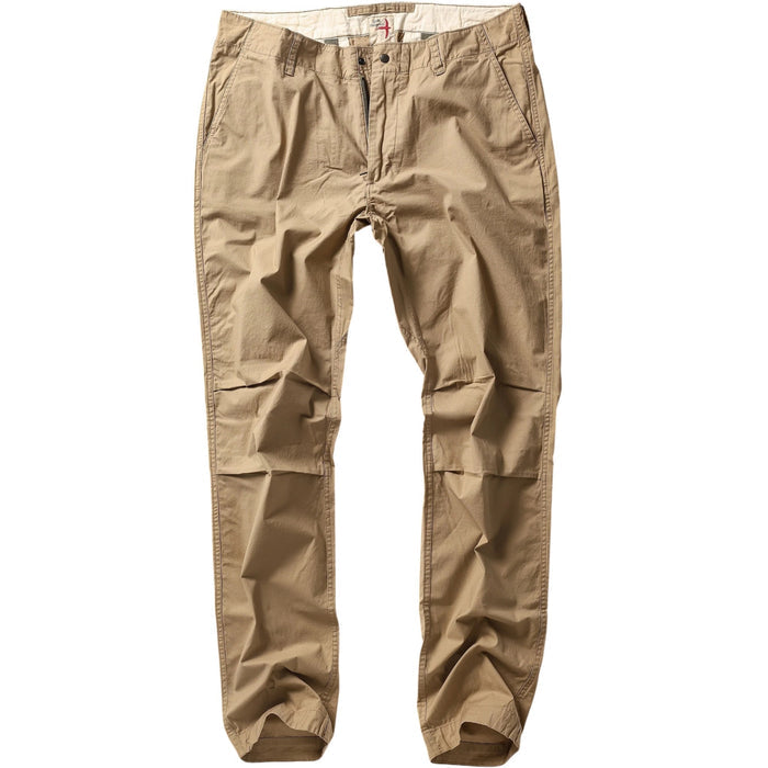 Wearing RELWEN's Flyweight Flex Chino in British Khaki, a person stands on a beachside boardwalk near a weathered bench, with flip-flops and a plaid shirt, seashells and sand in the background.