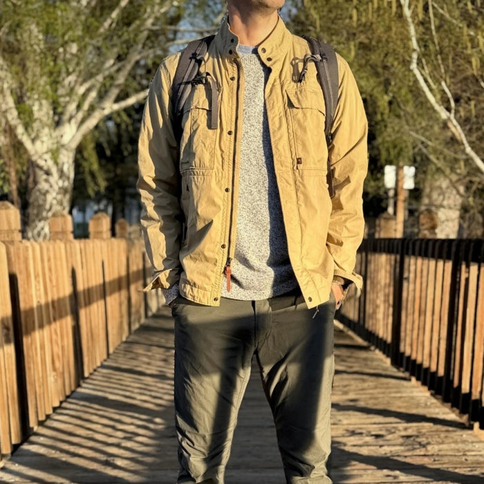 A person wearing the RELWEN Sailcloth Tanker in Desert Khaki stands on a wooden walkway, hands in pockets, with trees and sunlight in the background.