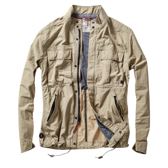 A person wearing the RELWEN Sailcloth Tanker in Desert Khaki stands on a wooden walkway, hands in pockets, with trees and sunlight in the background.