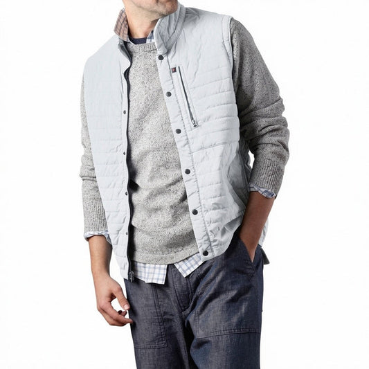 A person models the RELWEN - WINDZIP VEST IN GHOST GRAY over a gray sweater, plaid shirt, and dark blue jeans, standing with one hand in their pocket against a white background for extra layered warmth.