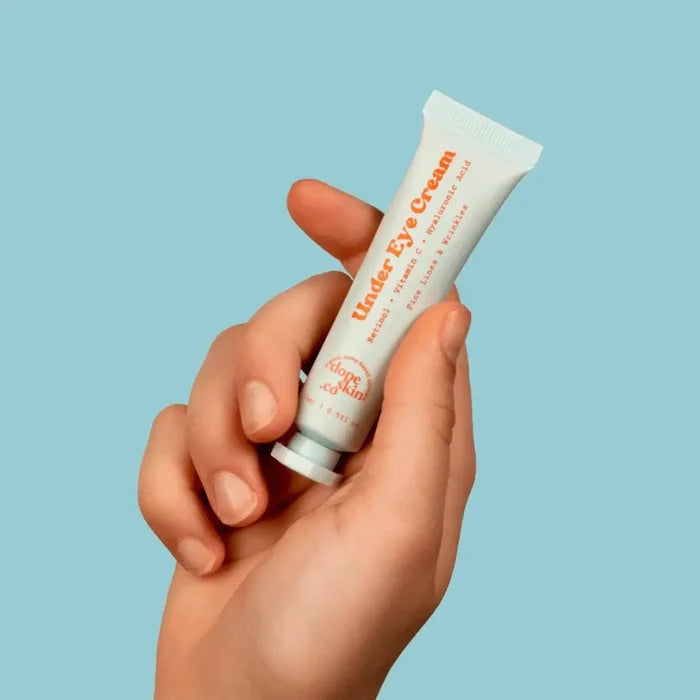 Hand holding a tube of Under Eye Cream against a light blue background