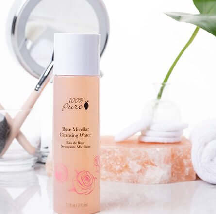 Bottle of 100% Pure Rose Micellar Cleansing Water on a bathroom counter with a mirror and towel in the background.