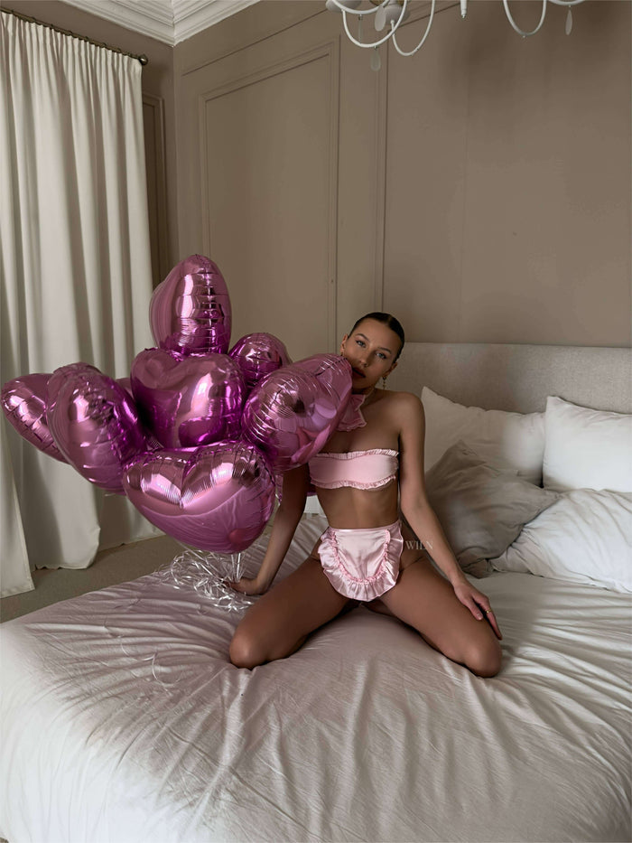 Model in pink SECRET 3 Piece Cosplay Lingerie Set surrounded by heart-shaped balloons on a bed.