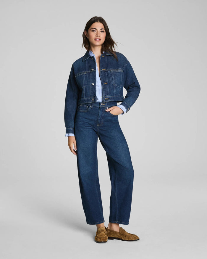 Here is the rewritten sentence using your product data: A woman with long dark hair and a relaxed expression stands against a plain background wearing the SPANX SPANXSHAPE Authentic 360 Barrel Leg Jean in Adriatic Blue, paired with a matching blue denim jacket, light blue shirt, and brown suede loafers.