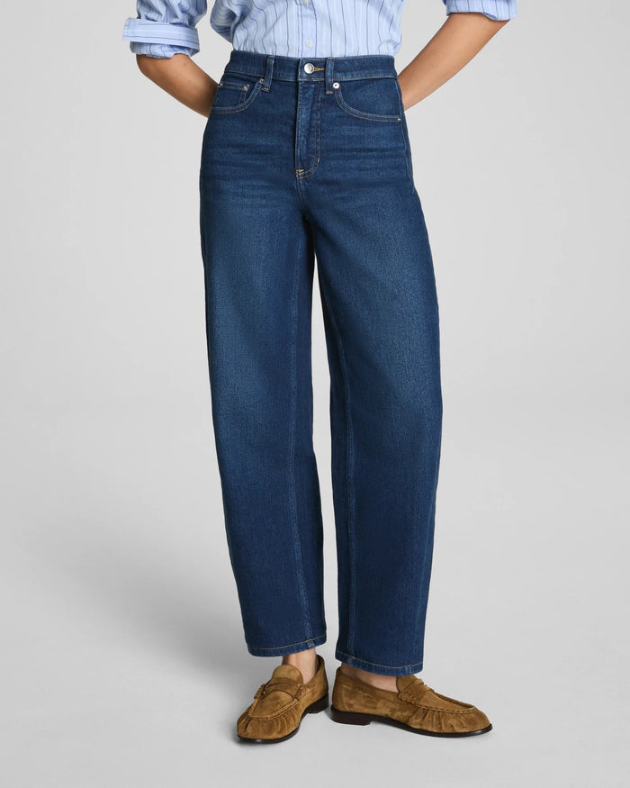 Here is the rewritten sentence using your product data: A woman with long dark hair and a relaxed expression stands against a plain background wearing the SPANX SPANXSHAPE Authentic 360 Barrel Leg Jean in Adriatic Blue, paired with a matching blue denim jacket, light blue shirt, and brown suede loafers.