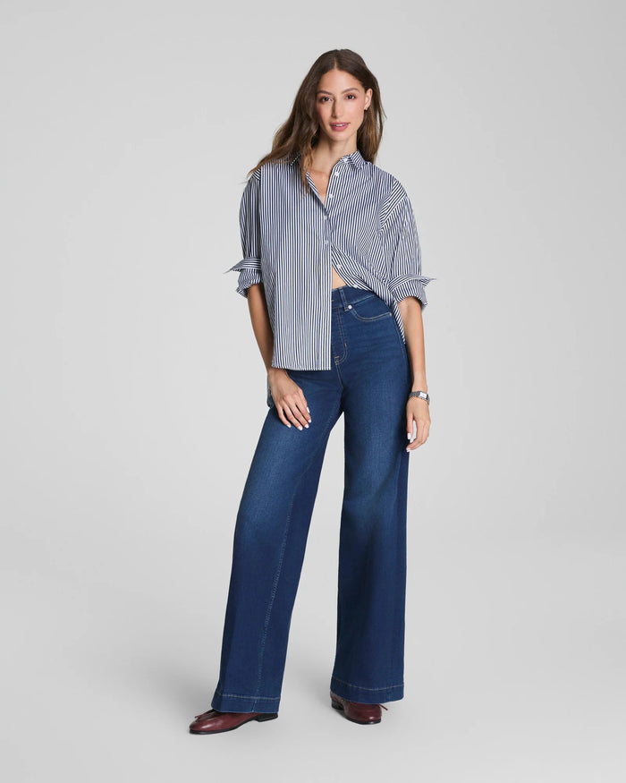 A woman with long brown hair stands against a plain background, smiling slightly. She is wearing a blue and white striped blouse paired with the SPANX Shape EveryWear Pull On Wide Leg Jean in Poseidon by SPANX and brown shoes, accessorized with a watch on her wrist.