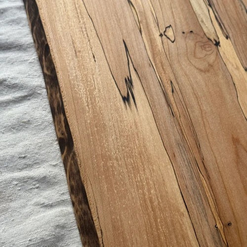 Handcrafted by PETERMANS BOARDS & BOWLS INC, the SPENCER PETERMAN - NATURAL EDGE SPALTED MAPLE RECTANGULAR BOARD 15" features a beautiful natural spalted maple grain, slightly angled sides, and a convenient small hole in the top right corner for hanging. Its dark rustic edges highlight the wood's unique character, making it an elegant choice for serving charcuterie or as a decorative cutting board.