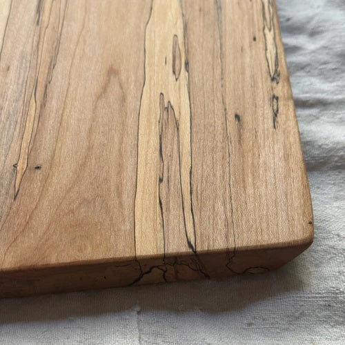 Handcrafted by PETERMANS BOARDS & BOWLS INC, the SPENCER PETERMAN - NATURAL EDGE SPALTED MAPLE RECTANGULAR BOARD 15" features a beautiful natural spalted maple grain, slightly angled sides, and a convenient small hole in the top right corner for hanging. Its dark rustic edges highlight the wood's unique character, making it an elegant choice for serving charcuterie or as a decorative cutting board.