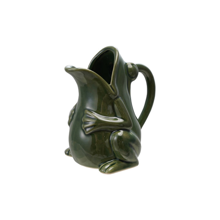 The CREATIVE COOP STONEWARE FROG PITCHER with a green reactive glaze sits on a wooden surface beside a potted variegated plant, against a plain light background.